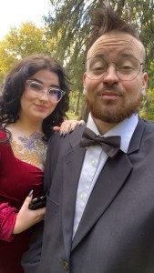 Kelsey black curly hair red velvet dress and Axton brown hair and suit selfie in front of the trees