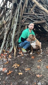 A man in jeans, a green shirt, and a backwards hat sits with his American Pitbull Terrier near a stick fort on a Morgan’s Knob loop. 
