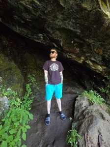 Axton in gray shirt, blue shorts and standing in a cave by the road.