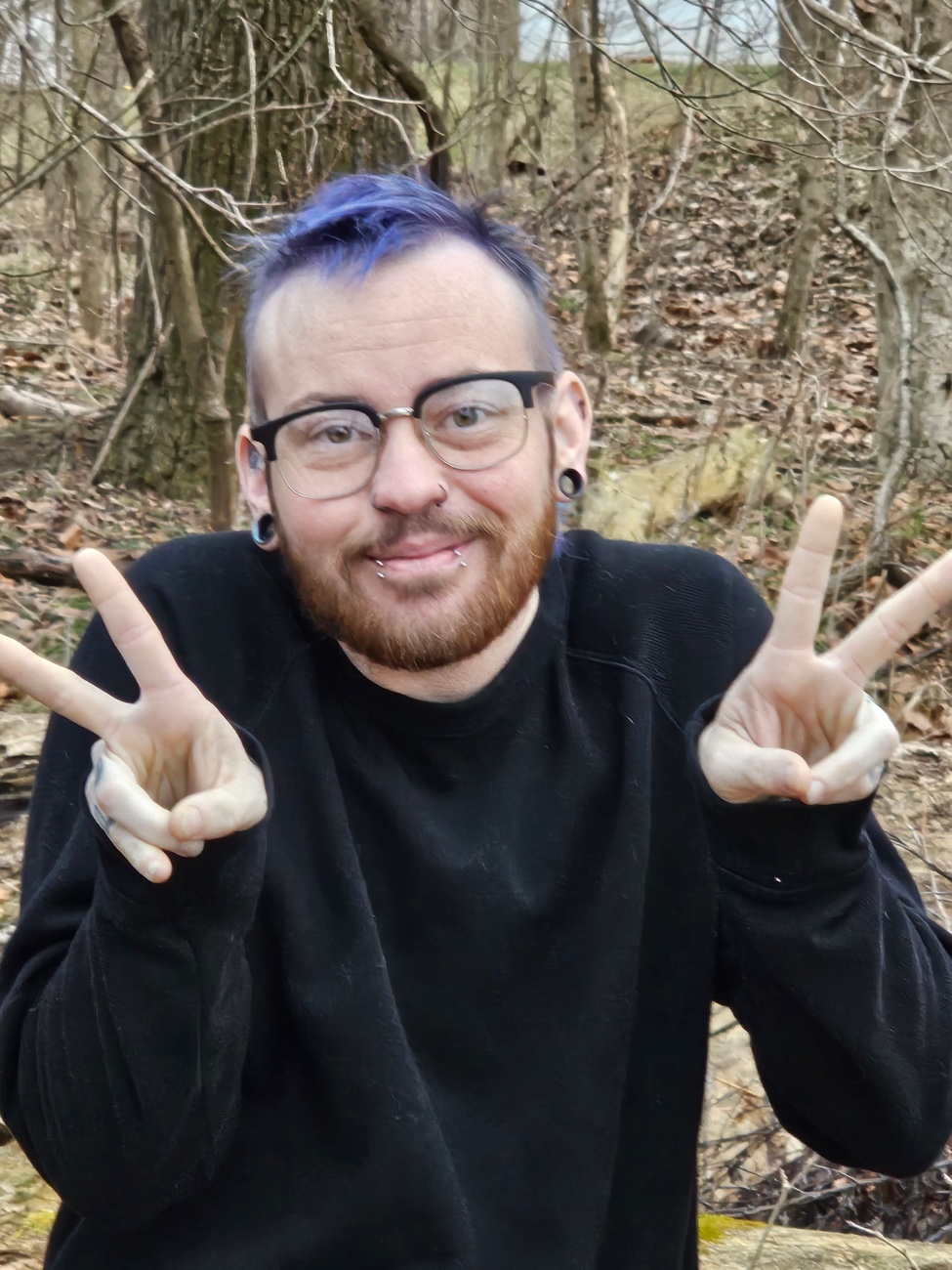 Axton with blue purple hair and a double peace sign in a black sweater in front of the winter woods.