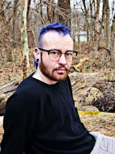 Axton Mitchell’s photographed in-front of bare winter trees purple mullet-hawk, glasses, and a black sweater. 