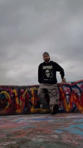 Axton leaning on a graffiti covered ledge in a black hoodie with a skull, gray joggers, a backwards hat, and green croc junipers at Mount Wood Overlook in Wheeling, WV