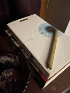 George Orwell's 1984 sitting on top of another book on a small stand next to an ashtray. A rolled marijuana cigar sits on the very top of the pile. 