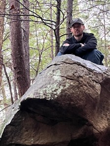 Axton in a black hat, black jacket , and jeans sitting on a large rock
