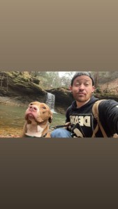Axton in a black hat, black jacket, and jeans with his dog in front of a waterfall