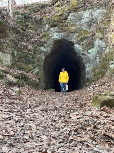 Axton in a Yellow jacket, black hat, and jeans stands in front of the ancient train tunnel