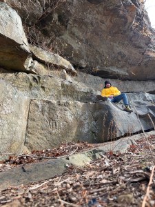Axton in a yellow jacket, black hat, and jeans sits on a cliff wall 