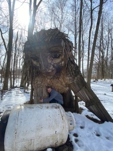 Axton in a grey jacket and jeans hugs the recycled wooden troll Bodil as they are surrounded by snow