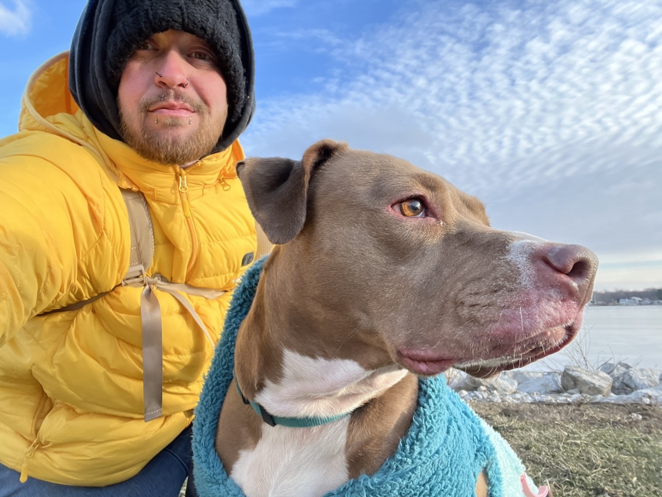 Axton in a yellow coat and black hat luna the rednosed pitbull in a blue jacket with blue sky and white clouds behind