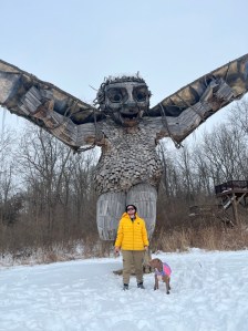 Luna in boots and a tie die coat with Axton in his yellow jacket in front of Bibbi the troll 