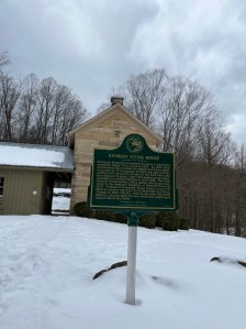 Snow surrounds the Kennedy stone house and green historical sign at salt fork