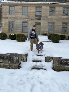 Luna in a blue shirt and Axton in tan khaki pants and a grey jacket outside the Kennedy stone house at saltfork surrounded by snow and green bushes