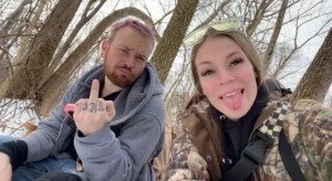 Axton and Hayley sit among the bare trees and snow. Axton in jeans and a grey jacket Hayley in a cameo jacket