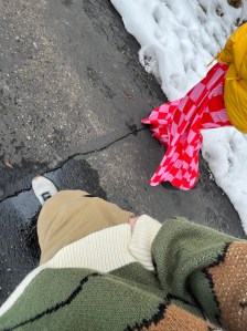 Axton wears Khaki pants, grey shoes, and a multi-color sweater viewed from the top looking down. Next to Kelsey in pink and red checkered print pants and a puffy yellow jacket. Not showing more than mid- torso down snow lines the asphalt trail
