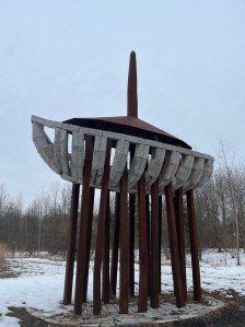 Granite steel and other recycled materials make up this grey and rust colored boat sculpture surrounded by bare winter trees and fallen snow 