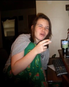 Axton is wearing Long dirty blonde hair, mtn dew pants, gray shirt and smoking a cigarette 