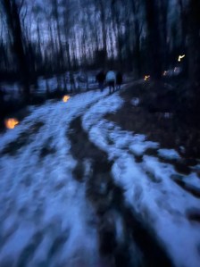 Milk jugs with flameless candles line the melting snow patchy trail, the brown colored ground shows through in the melted snow, bare trees, right before twilight 
