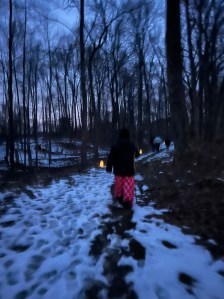 Snow melts slowly some brown ground shows through, an individual in a black coat, with black hair, and pink and red checkered pants walks into the barren tree, woods.
