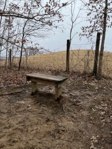 A wooden bench surrounded by melting snow, winter fields, bare trees, and mud. 