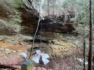 Semi-frozen waterfall dripping onto large ice formation surrounded by sandstone cliffs and bare trees