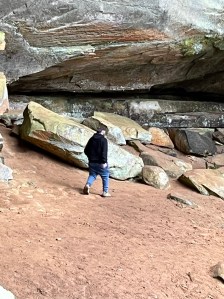 Axton walking under the cliffs near the waterfall, wearing jeans and a black hoodie, surrounded by red dirt and large rocks