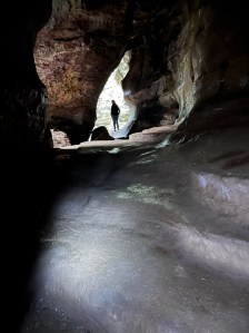 Axton in blue jeans black hoodie and hat stands in window to rock house in hocking hills surrounded by orange and red sandstone