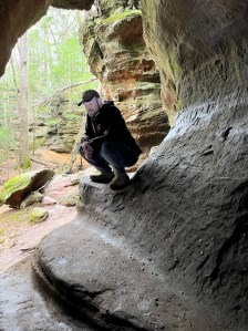 Axton in black hat and hoodie with blue jeans crouches in cave window