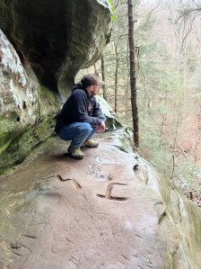 Axton crouching on cliff ledge wearing black hat and hoodie with blue jeans